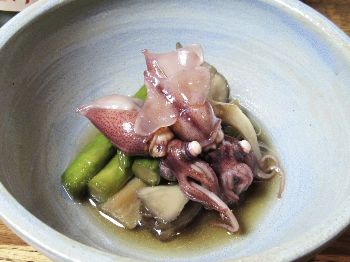 A bowl of Japanese-style simmered baby squid served with asparagus and mushrooms in a light soy-based broth, presented in a rustic ceramic dish.