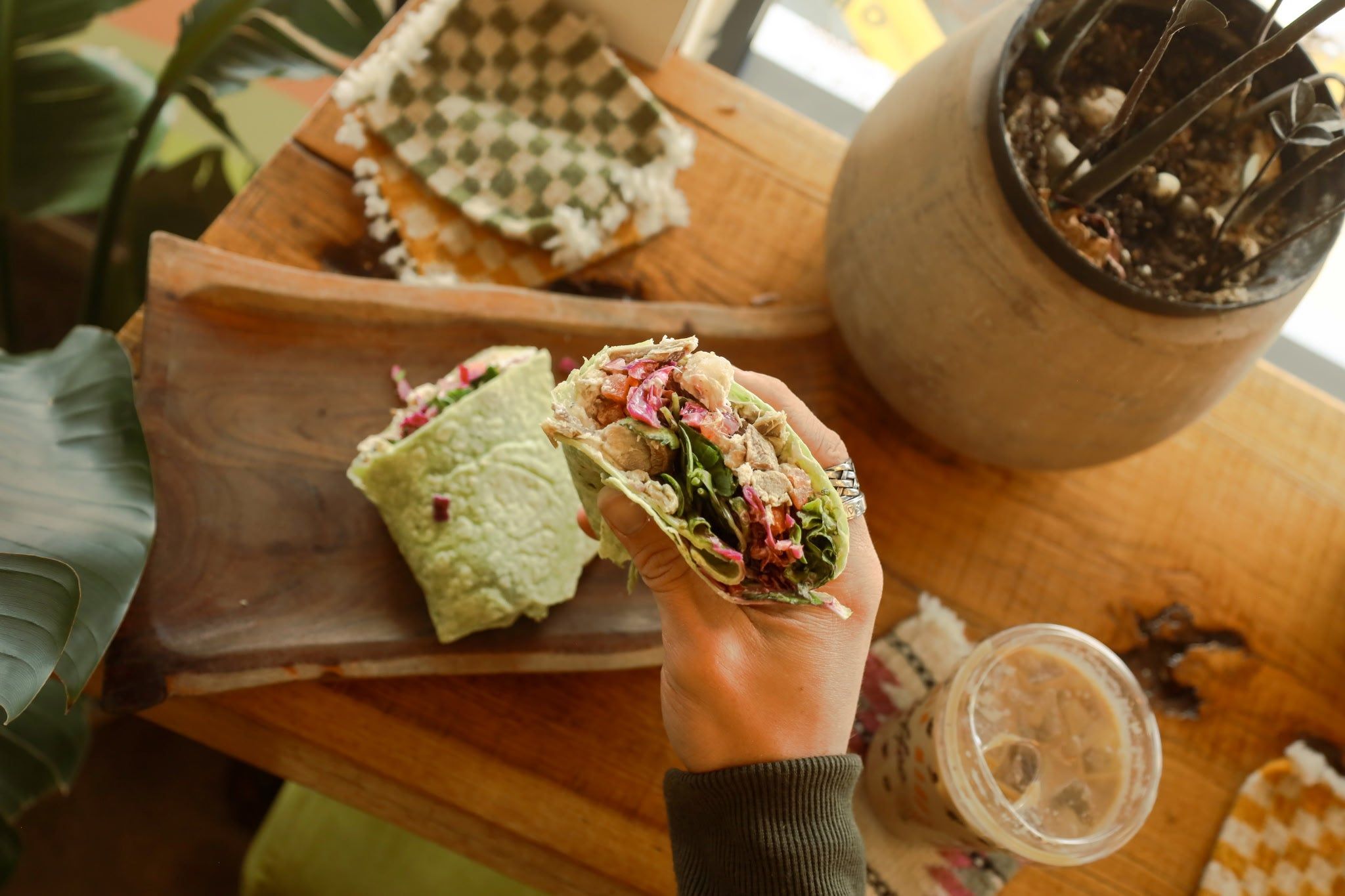 A hand holding a fresh wrap filled with colorful greens, grilled chicken, and veggies, paired with iced coffee. The cozy wooden counter and natural light create a relaxed small-town coffee shop vibe.