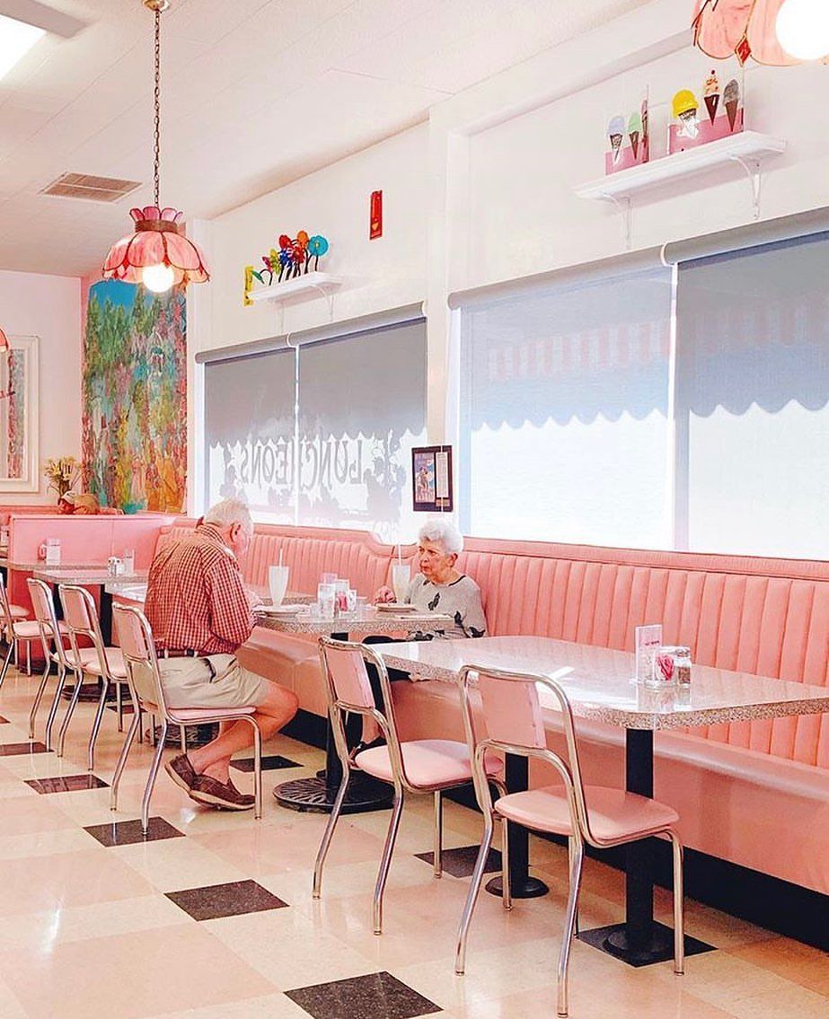 An elderly couple enjoys milkshakes in a retro pink diner with vintage decor and pastel booths. The nostalgic scene feels timeless, warm, and full of sweet charm.