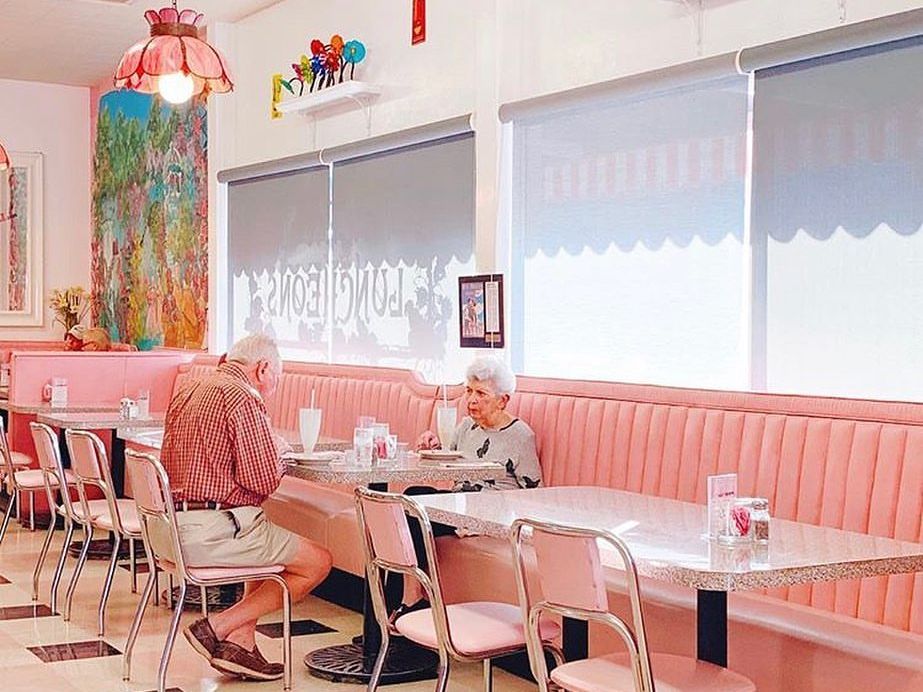 An elderly couple enjoys milkshakes in a retro pink diner with vintage decor and pastel booths. The nostalgic scene feels timeless, warm, and full of sweet charm.
