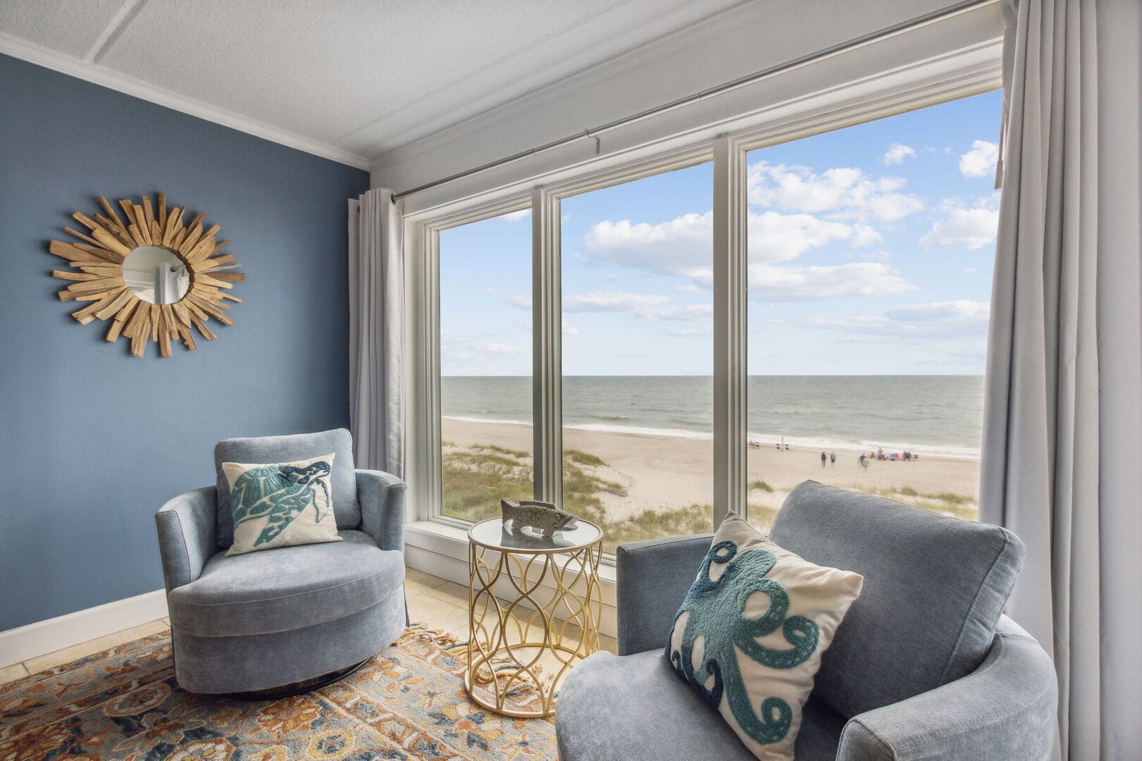 Cozy beachfront sitting area with two blue chairs, sea-themed pillows, and a gold side table, overlooking the ocean through large windows
