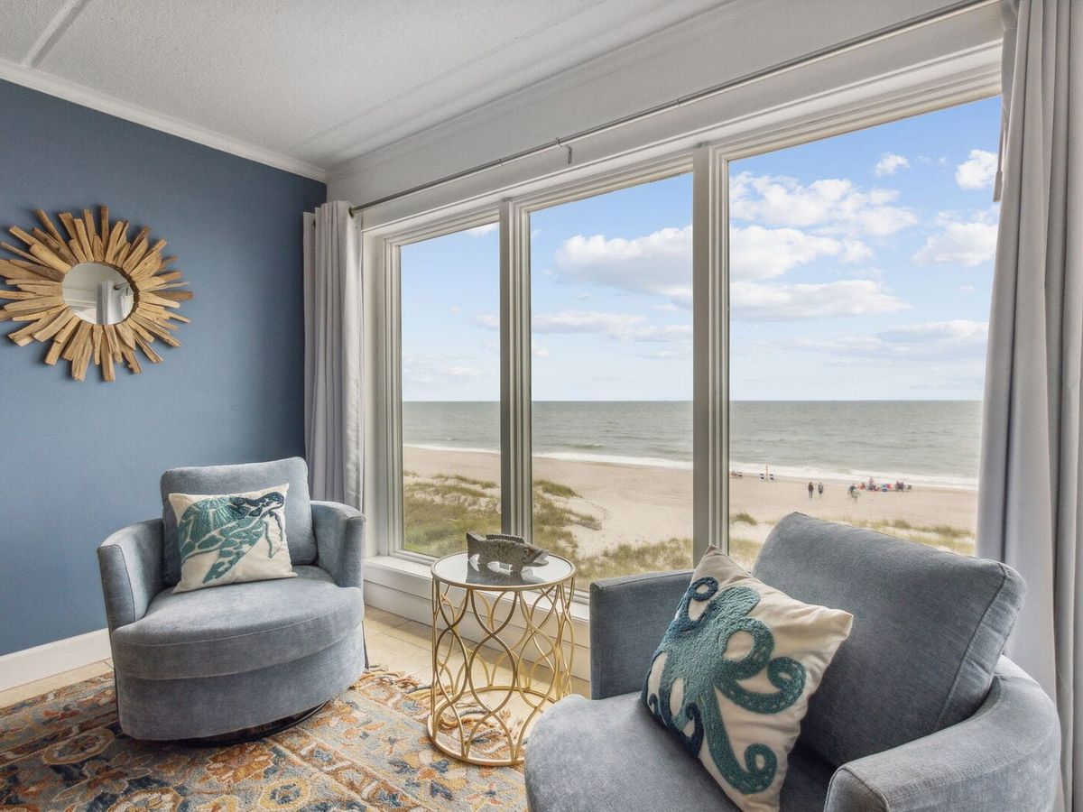 Cozy beachfront sitting area with two blue chairs, sea-themed pillows, and a gold side table, overlooking the ocean through large windows