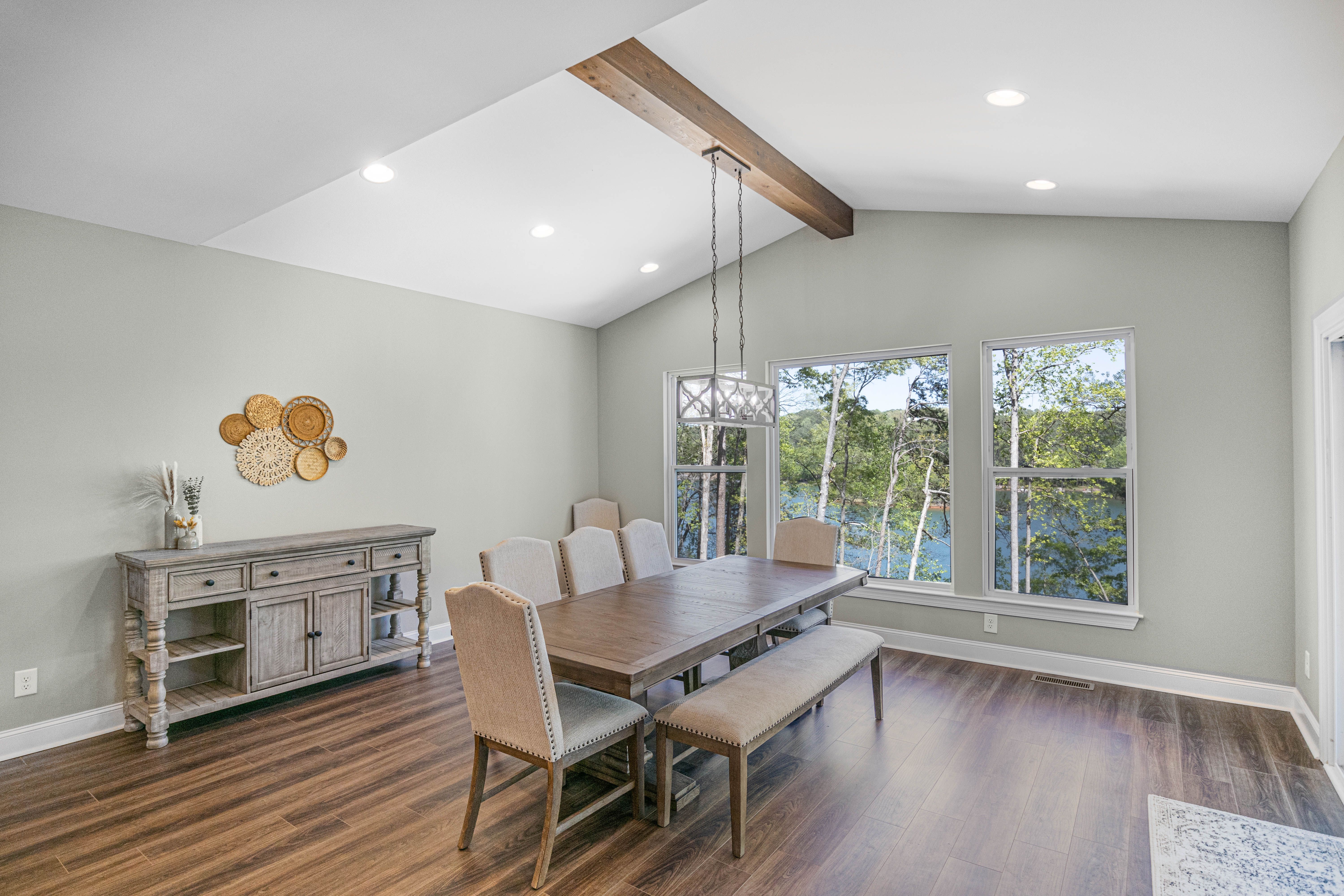 Dining room with vaulted ceiling, wood beam accent, modern chandelier, wooden table with upholstered chairs and bench seating, sideboard cabinet, and large windows overlooking trees and a lake.
