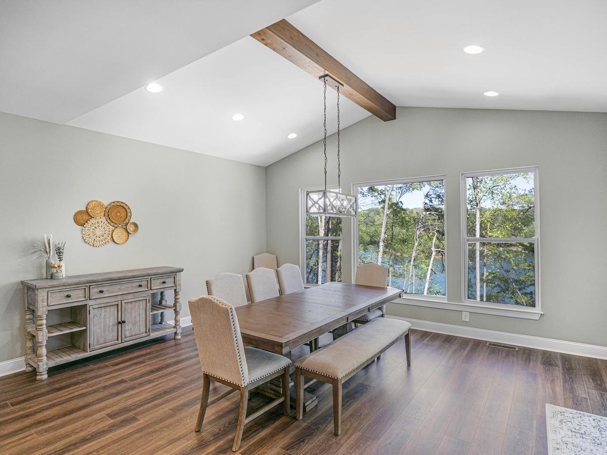 Dining room with vaulted ceiling, wood beam accent, modern chandelier, wooden table with upholstered chairs and bench seating, sideboard cabinet, and large windows overlooking trees and a lake.