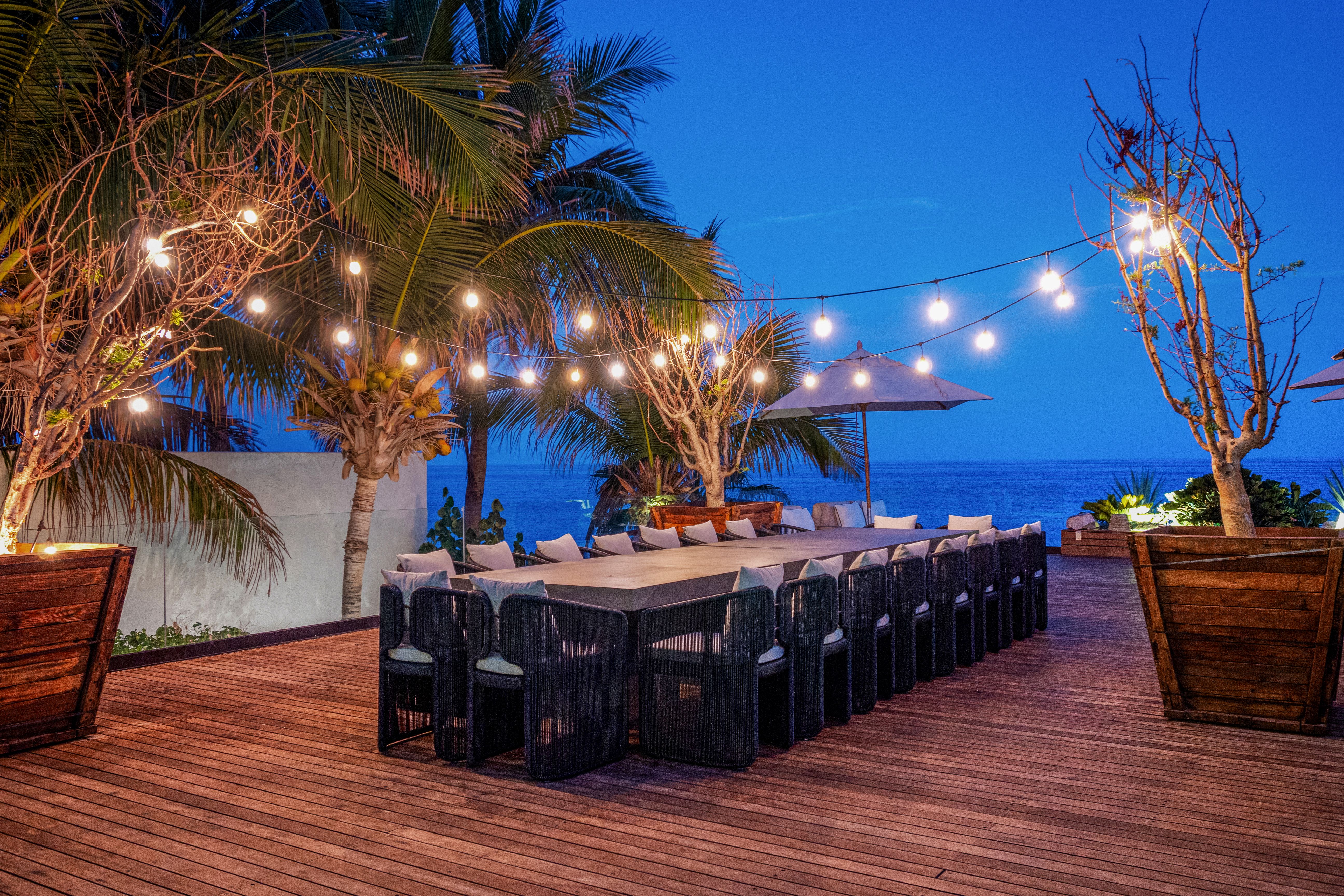 Oceanfront dining deck at twilight with a long table, cushioned chairs, string lights overhead, and palm trees framing the sea view.