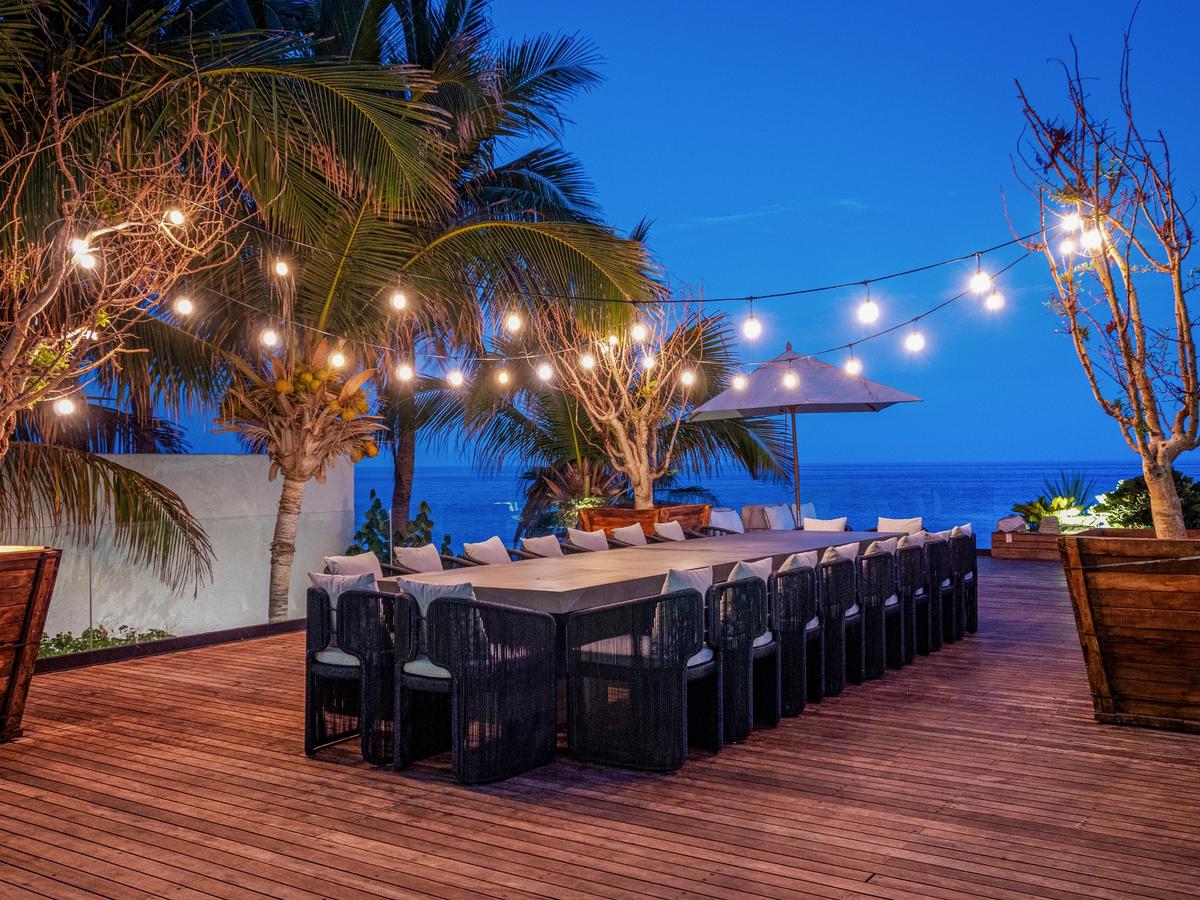 Oceanfront dining deck at twilight with a long table, cushioned chairs, string lights overhead, and palm trees framing the sea view.