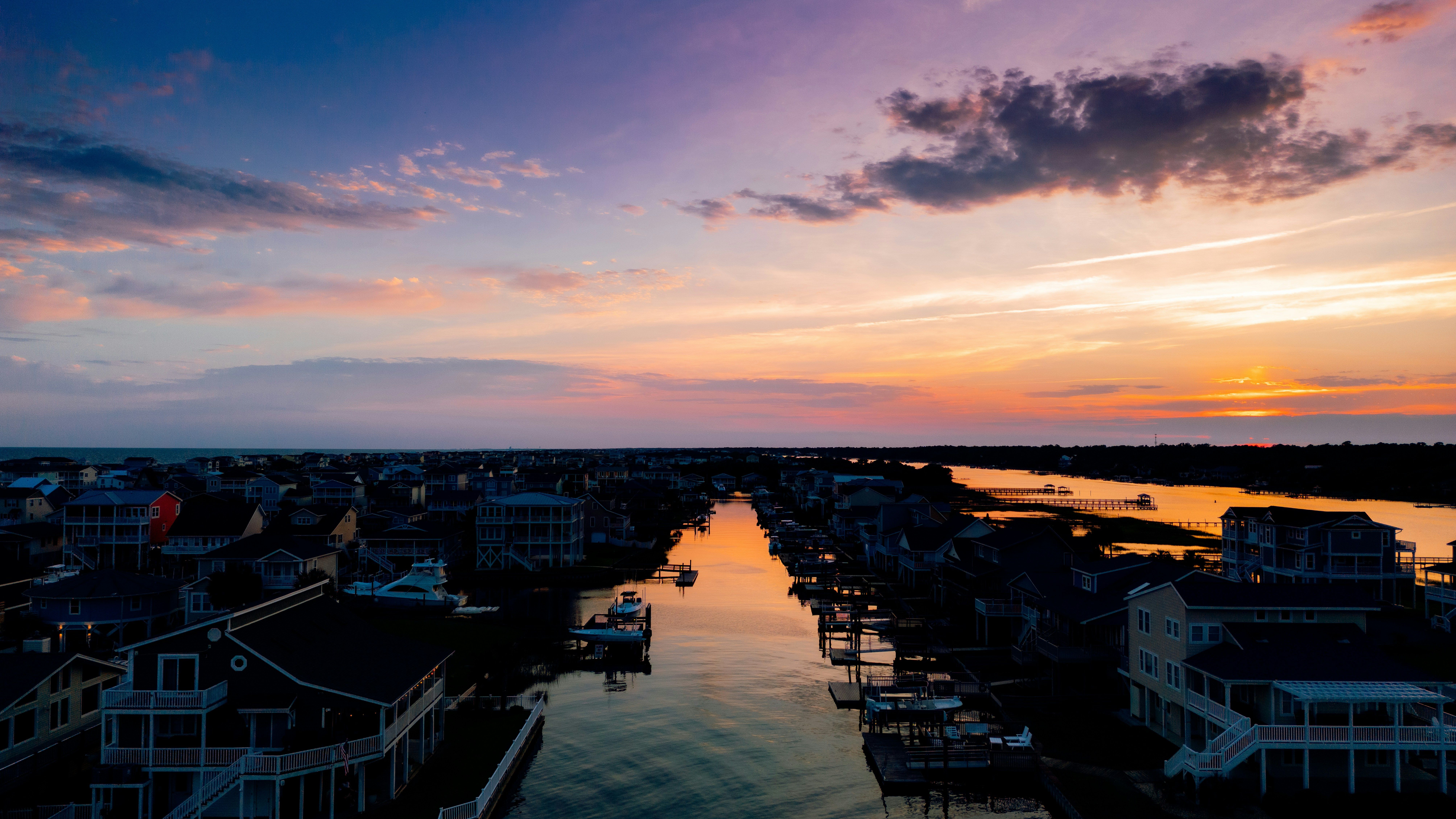 Aerial view of a coastal canal community at sunset, with waterfront homes and docks reflecting the orange and purple hues of the sky over calm waters.