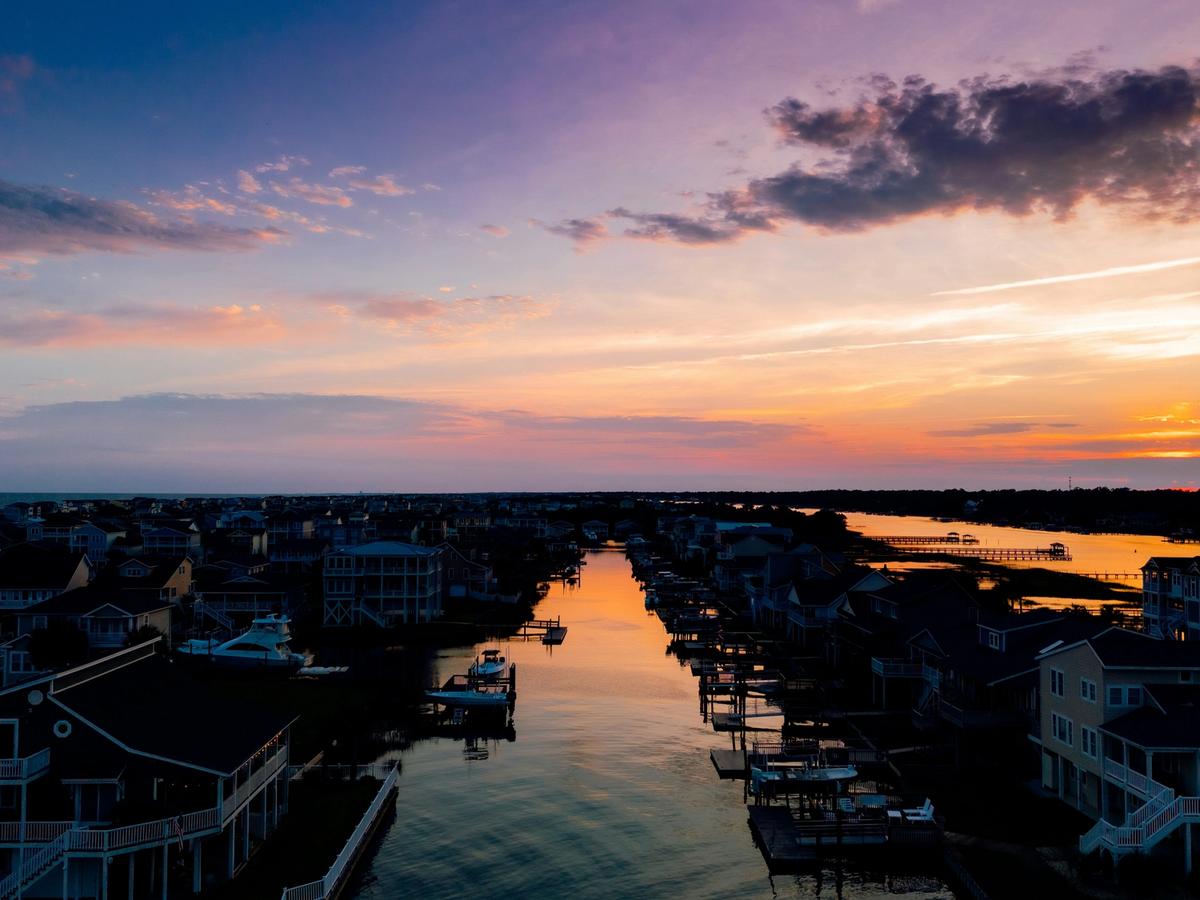Aerial view of a coastal canal community at sunset, with waterfront homes and docks reflecting the orange and purple hues of the sky over calm waters.