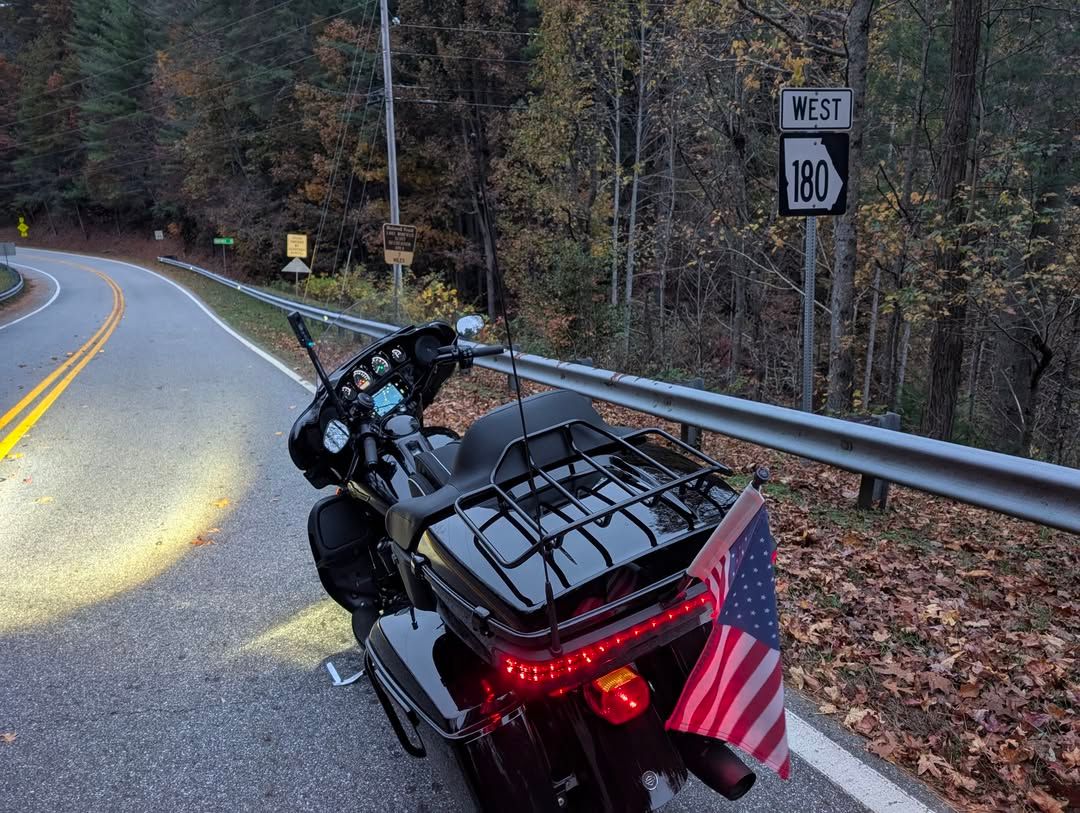A motorcycle with an American flag is parked along a winding mountain road lined with autumn trees. The quiet curve of Georgia Route 180 sets the scene for a perfect scenic ride through the hills.