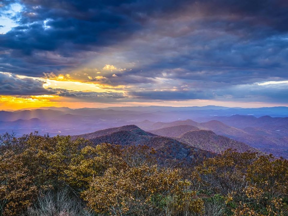 Golden sunset light breaks through the clouds over the rolling mountain ridges at Brasstown Bald, the highest point in Georgia. This scenic overlook offers wide views of the Blue Ridge Mountains, making it a popular spot for sunrise and sunset photos in the fall.