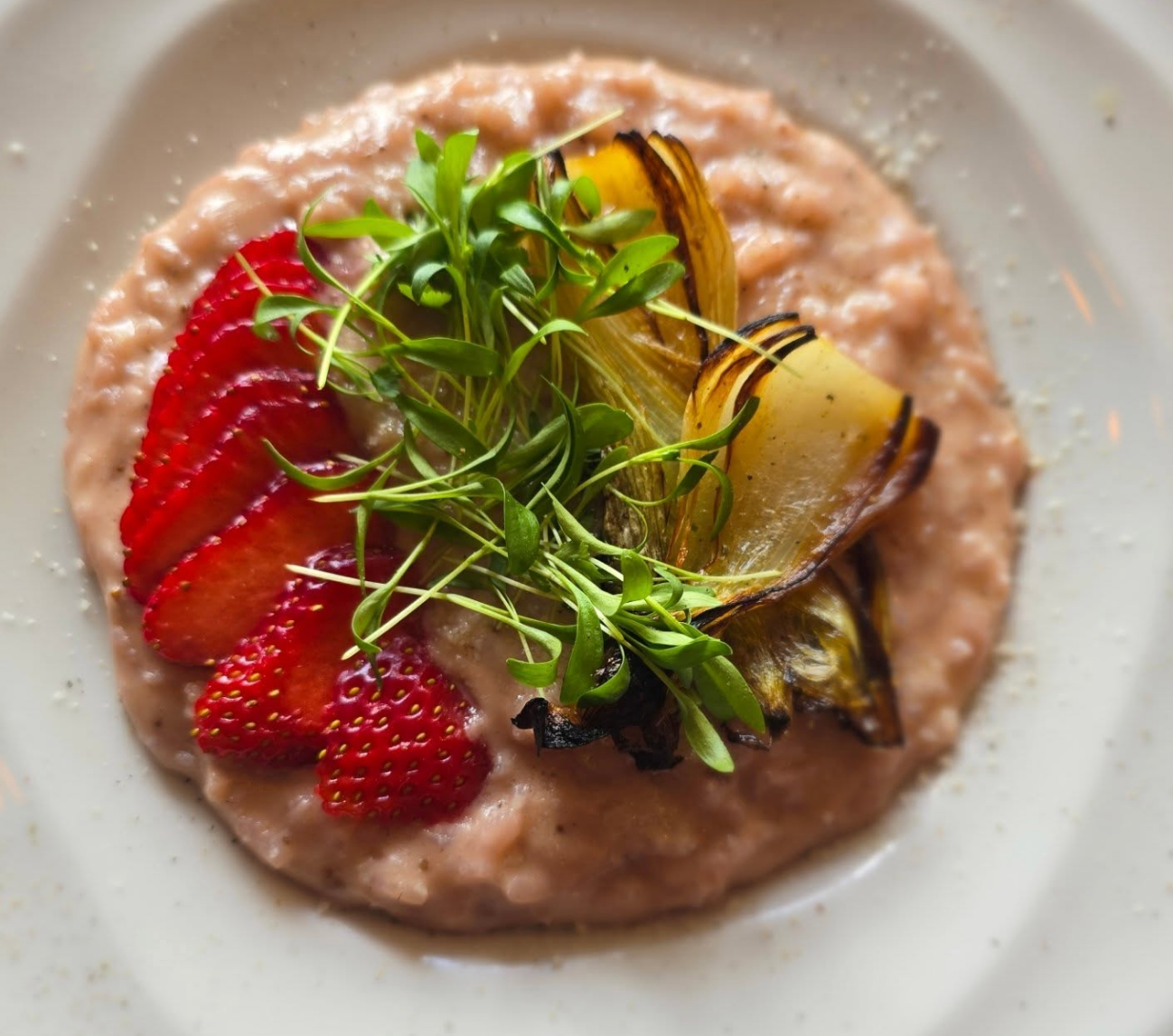 A gourmet plate of creamy pink risotto topped with sliced strawberries, charred endive, and fresh microgreens.