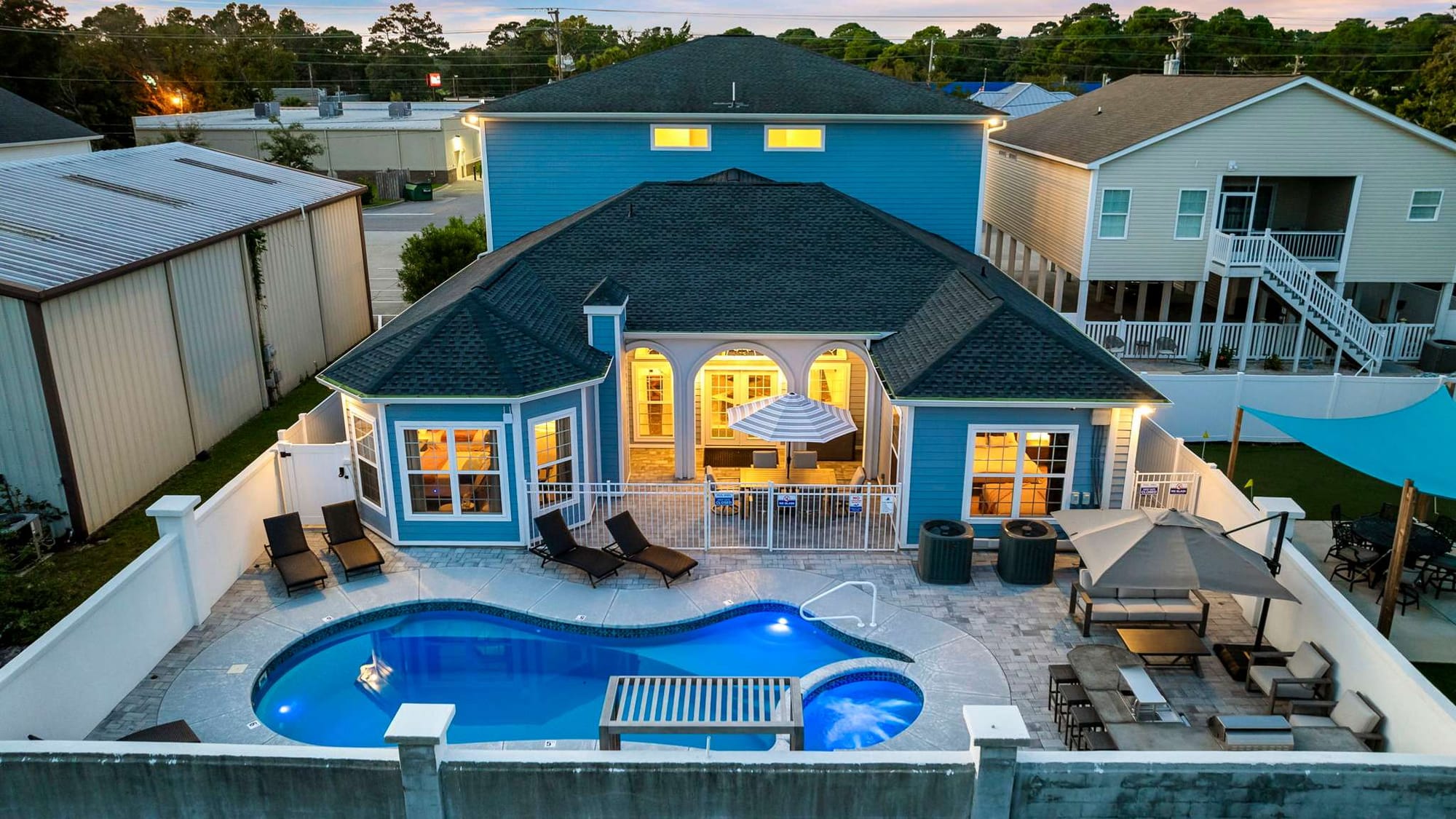 This bright blue beach retreat offers a private pool, spacious patio, and resort-style lounge chairs—ideal for large family vacations in North Myrtle Beach. Inside, wide windows glow warmly at dusk, creating a relaxing coastal vibe.