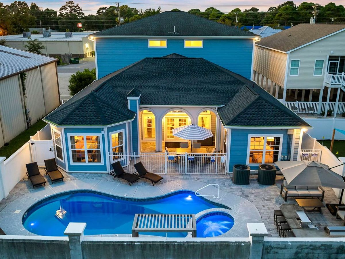 This bright blue beach retreat offers a private pool, spacious patio, and resort-style lounge chairs—ideal for large family vacations in North Myrtle Beach. Inside, wide windows glow warmly at dusk, creating a relaxing coastal vibe.