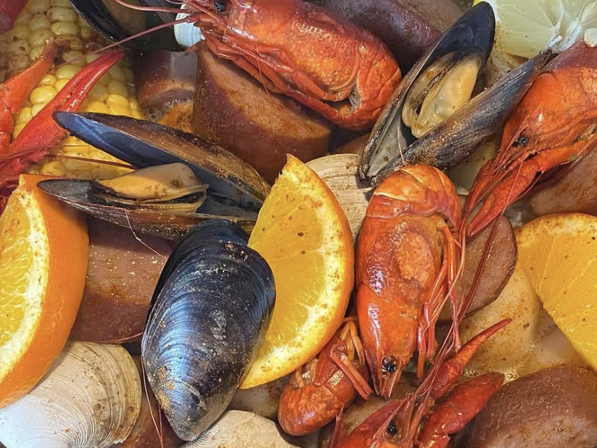 A close-up of a seafood boil featuring crawfish, mussels, clams, sausage, corn, and orange slices seasoned with spices.