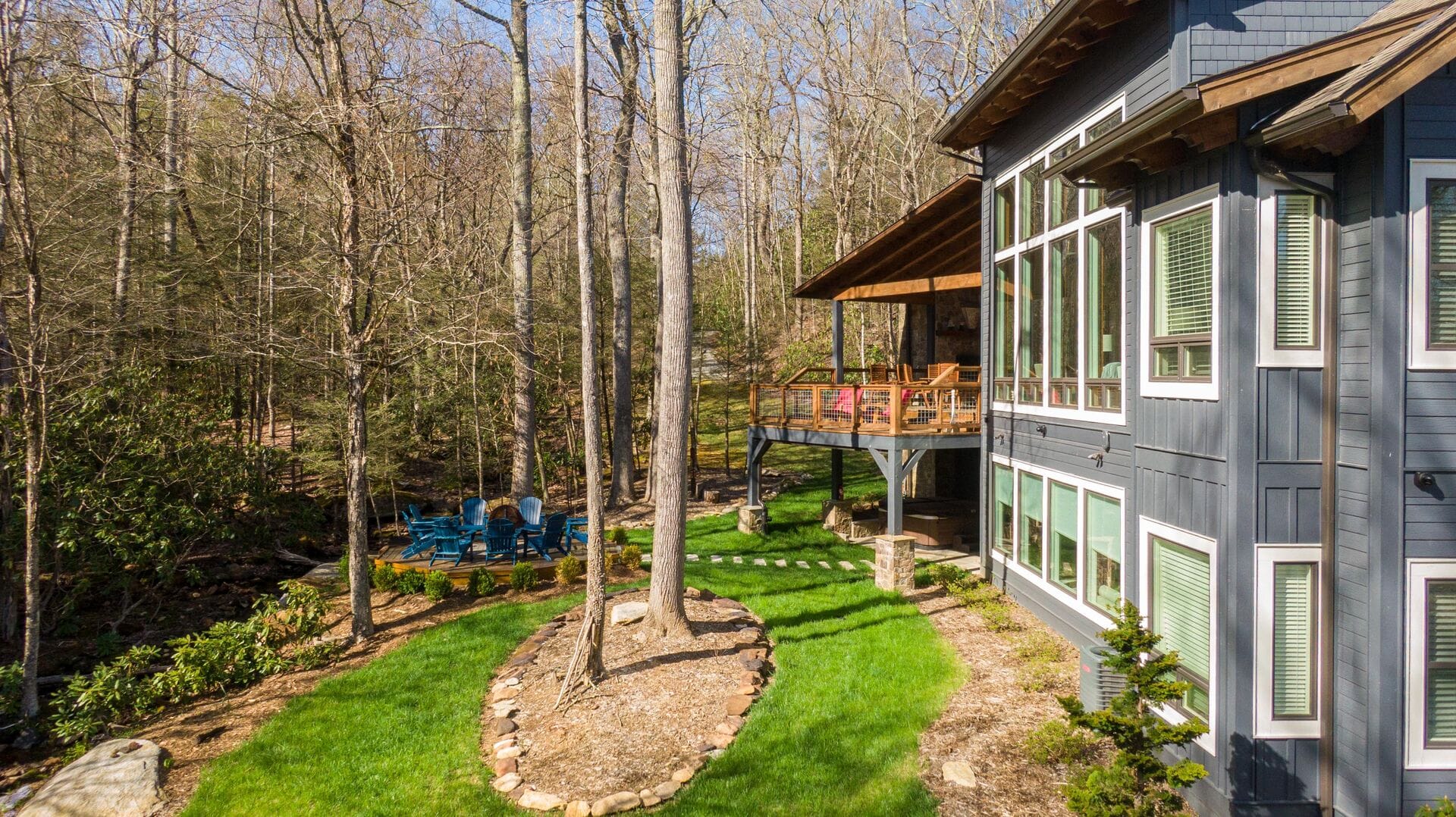 A cozy modern cabin with tall windows and a wraparound deck sits among tall trees in a forest clearing. Blue chairs circle a fire pit nearby, inviting guests to relax and enjoy the peaceful woods.