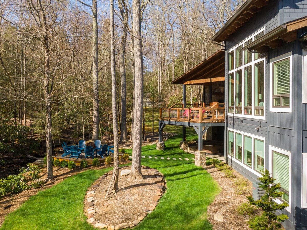 A cozy modern cabin with tall windows and a wraparound deck sits among tall trees in a forest clearing. Blue chairs circle a fire pit nearby, inviting guests to relax and enjoy the peaceful woods.