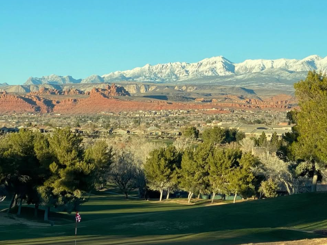 Picturesque golf course with lush green fairways, framed by towering trees, red rock formations, and snow-capped mountains under a clear blue sky