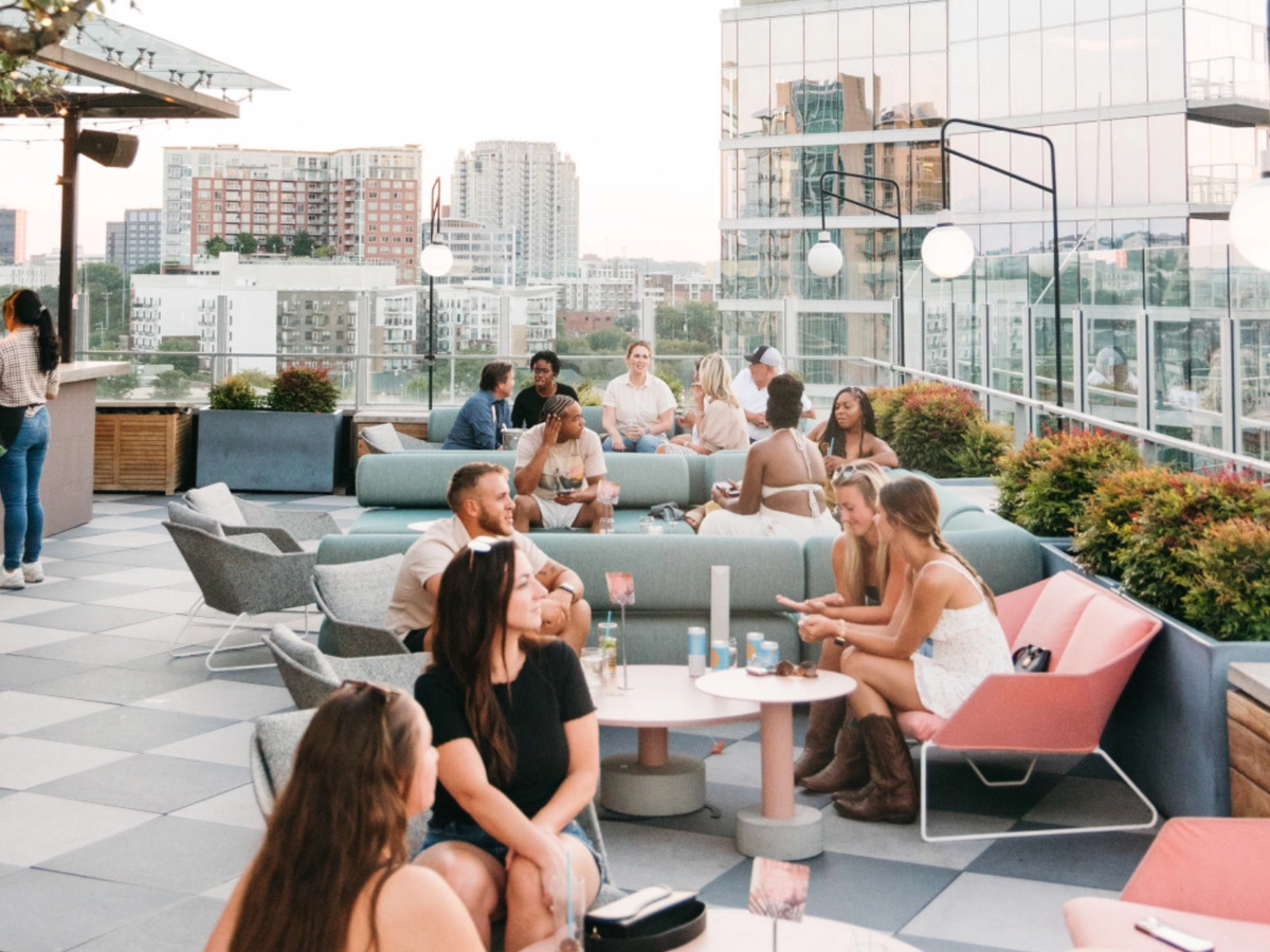 People relaxing and socializing on a stylish rooftop bar with modern seating, city views, and evening light reflecting off surrounding buildings.
