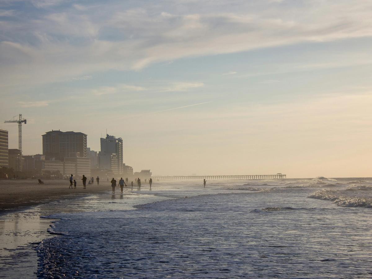 eople stroll along the Myrtle Beach shoreline at low tide as gentle waves roll in, with the pier and oceanfront skyline fading into soft, golden evening light.