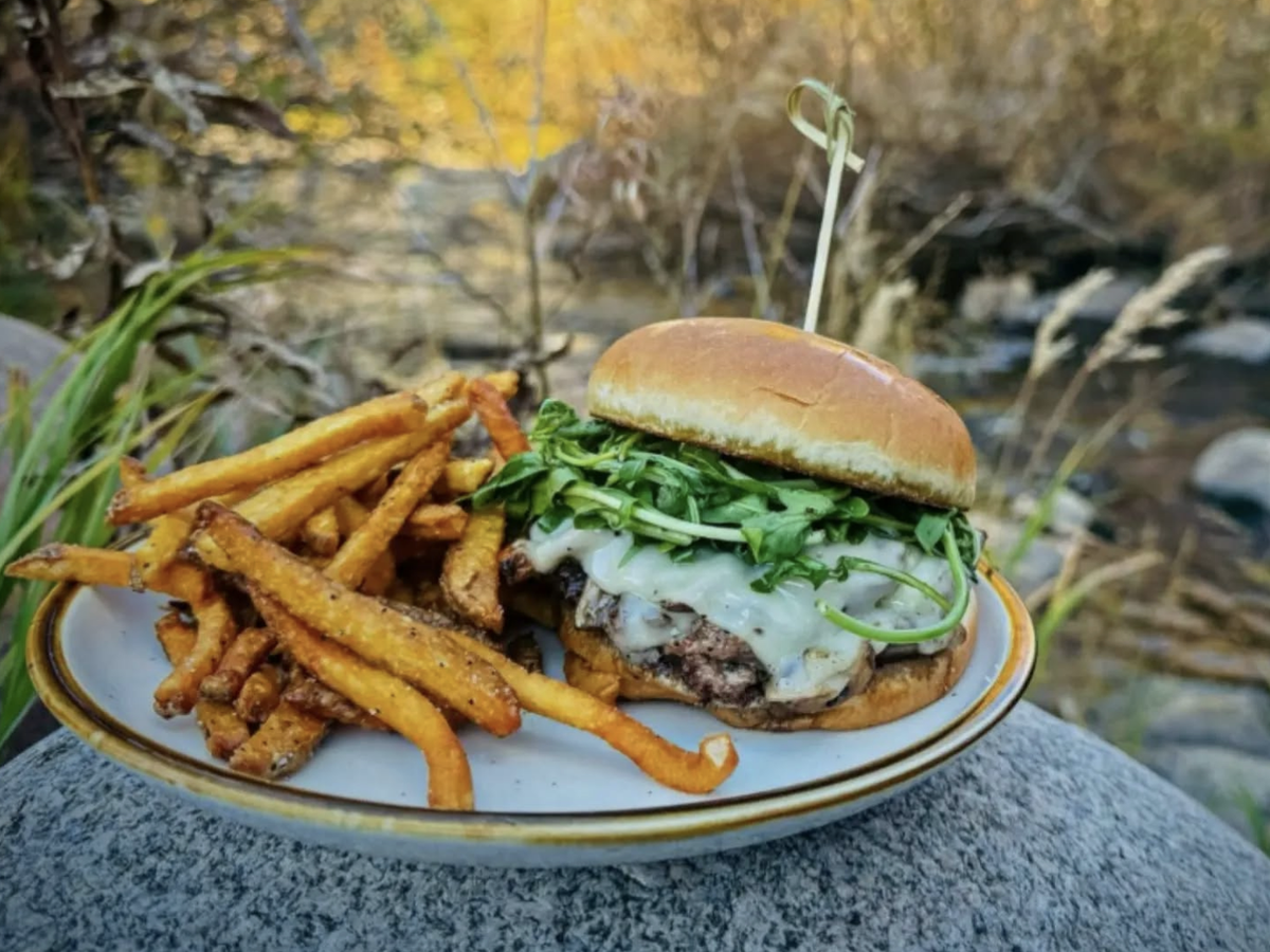 Juicy cheeseburger topped with arugula served with seasoned fries on a plate, set outdoors on a rock beside a stream and surrounded by autumn foliage.