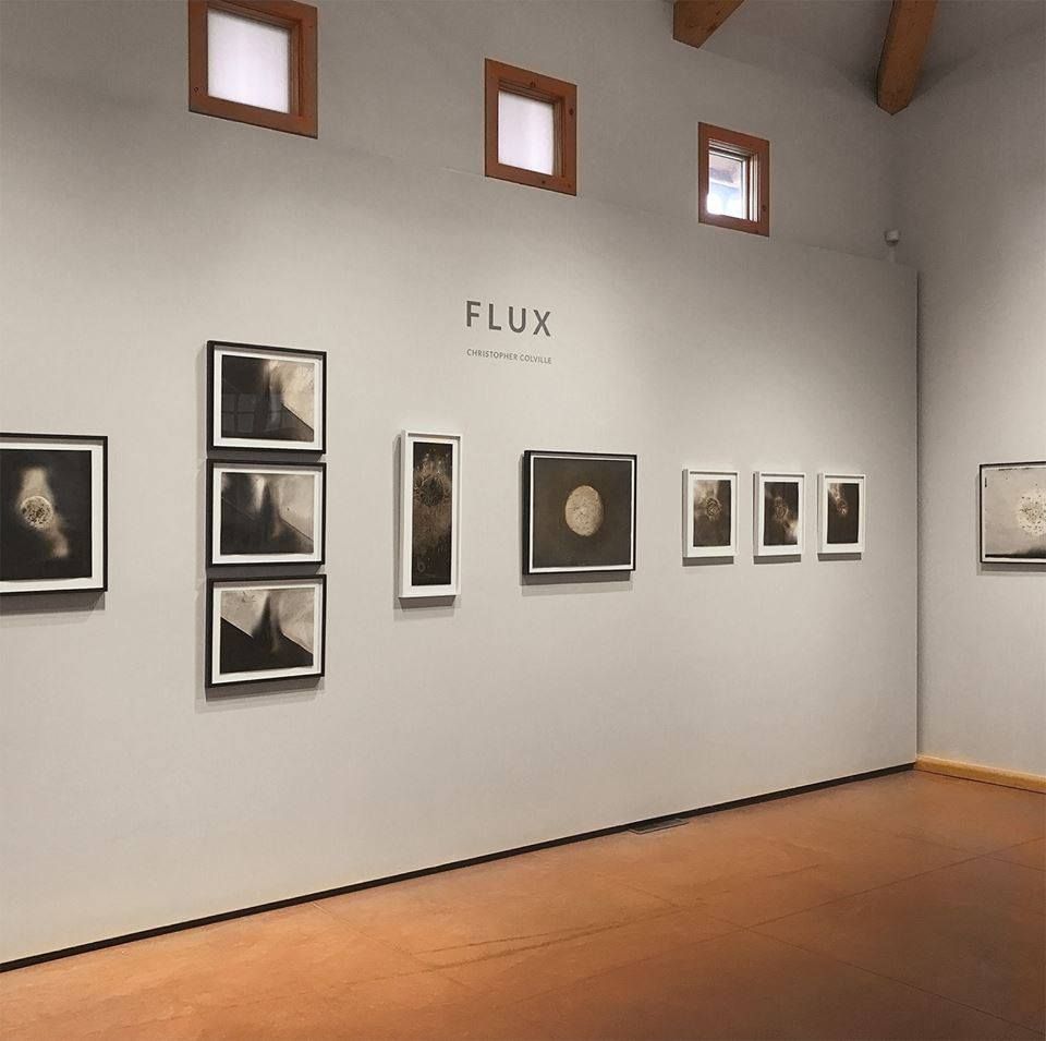 A calm modern gallery displays a series of dark-toned abstract artworks arranged neatly along a clean white wall. Soft natural light from the small windows above gives the room a warm and peaceful feeling. The minimalist setup lets the textures and details of each piece stand out.