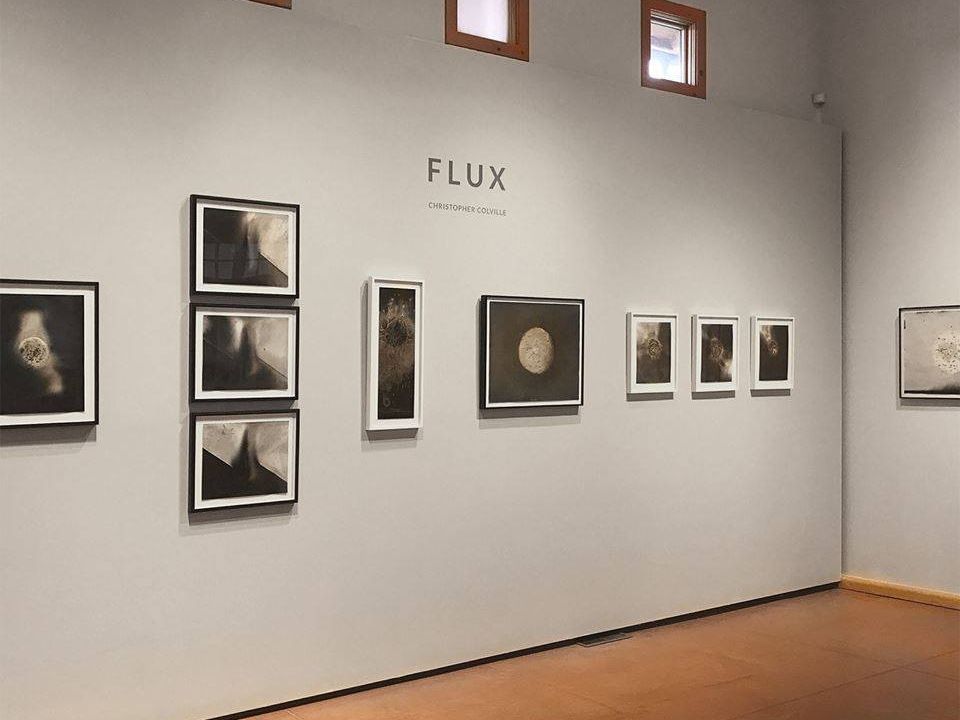 A calm modern gallery displays a series of dark-toned abstract artworks arranged neatly along a clean white wall. Soft natural light from the small windows above gives the room a warm and peaceful feeling. The minimalist setup lets the textures and details of each piece stand out.