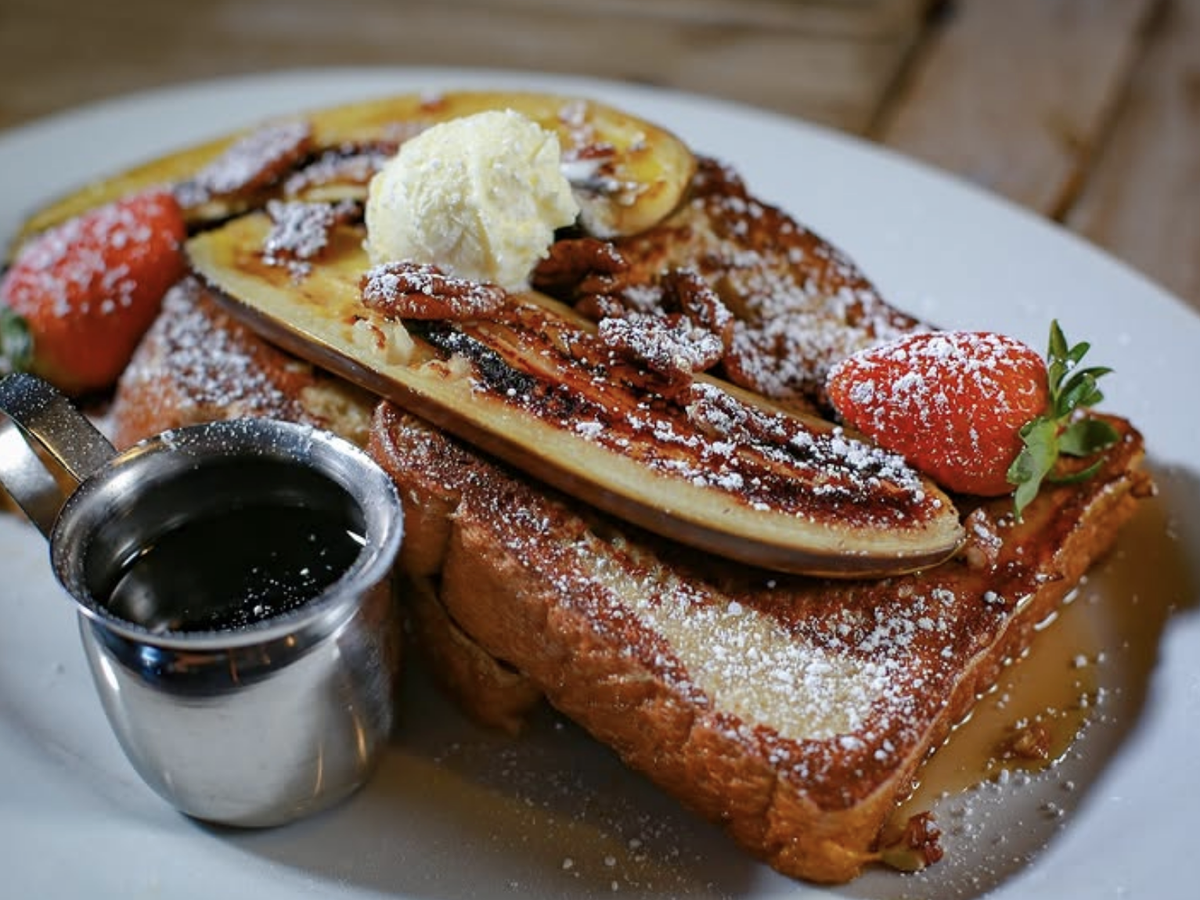 A plate of French toast topped with grilled bananas, pecans, a scoop of butter, and fresh strawberries, dusted with powdered sugar and served with a small metal cup of syrup.