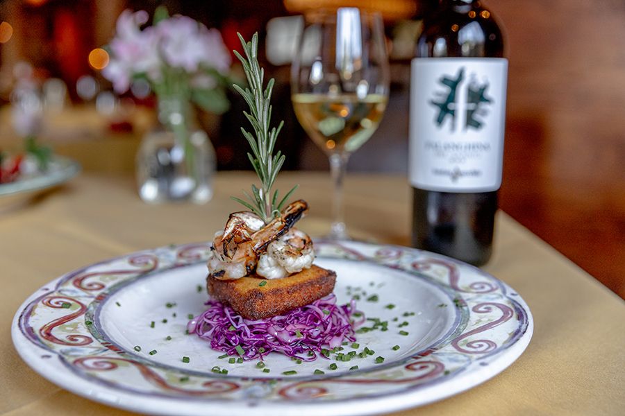 A beautifully plated dish of grilled shrimp, crispy polenta, and red cabbage sits on an elegant table beside a glass of white wine. The cozy restaurant atmosphere invites a perfect date night.