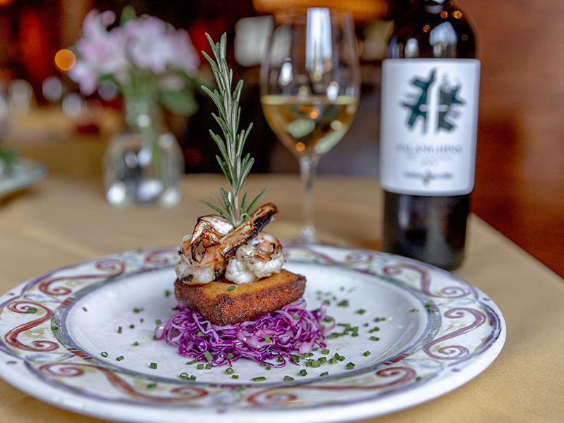 A beautifully plated dish of grilled shrimp, crispy polenta, and red cabbage sits on an elegant table beside a glass of white wine. The cozy restaurant atmosphere invites a perfect date night.