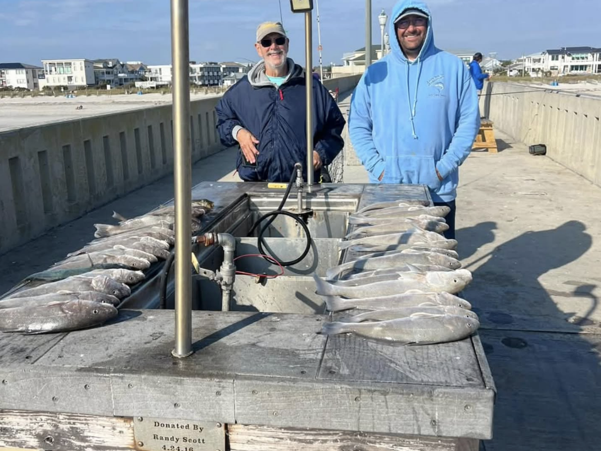 Two men standing at a pier cleaning station with a large display of freshly caught fish lined up on both sides of the table, with beach houses and ocean visible in the background.