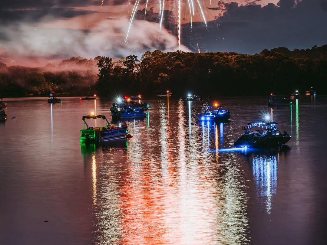 A scenic lakeside trail with boats and fireworks lighting up the evening sky, perfect for a sunset hike with a view. Hikers rest by the water, soaking in the festive atmosphere.