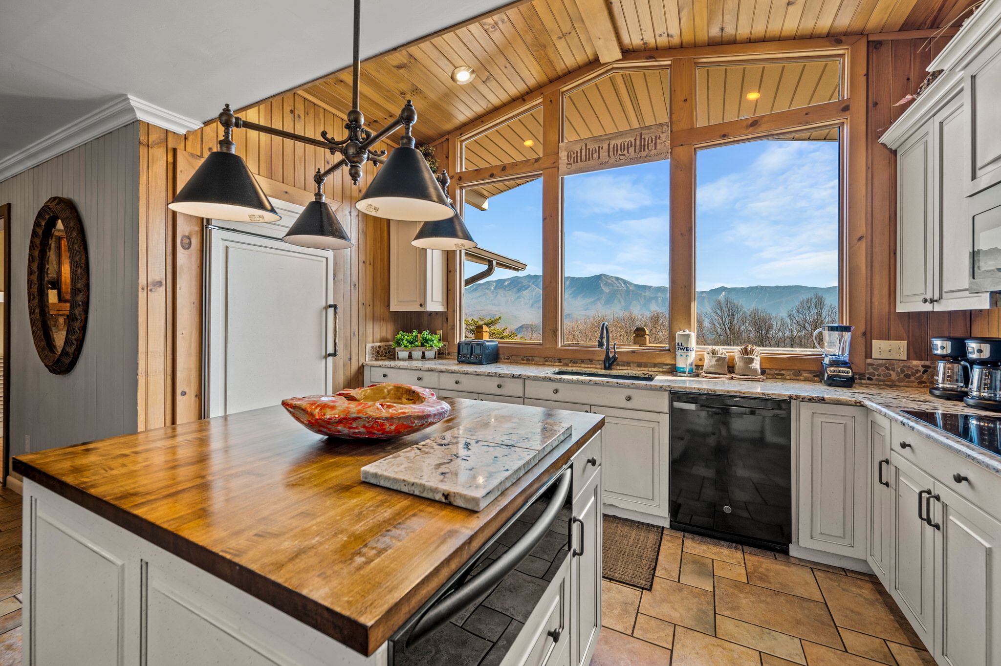 A rustic kitchen with a wooden ceiling and large picture windows showcasing mountain views, featuring white cabinetry, a wood and stone island, and black appliances with warm, cozy lighting.