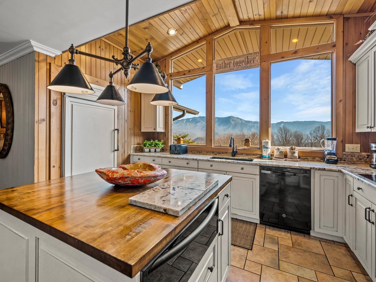 A rustic kitchen with a wooden ceiling and large picture windows showcasing mountain views, featuring white cabinetry, a wood and stone island, and black appliances with warm, cozy lighting.