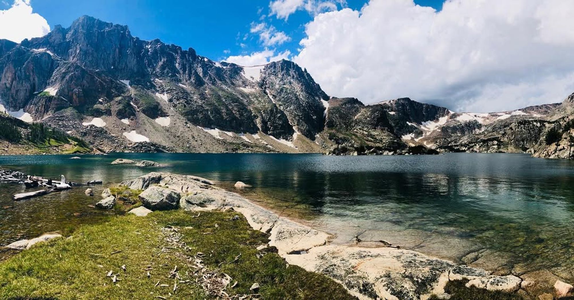 A stunning alpine lake with clear water reflecting jagged mountain peaks and patches of snow. A perfect summer hike with jaw-dropping views in Montana’s high country.