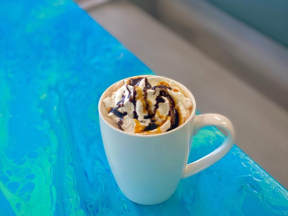 A white mug brimming with whipped cream, chocolate, and caramel drizzle sits on a vibrant blue swirled table.