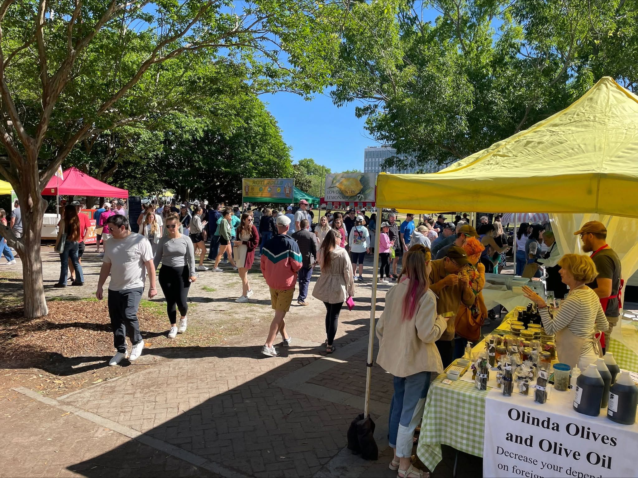 A lively crowd of shoppers browses colorful vendor tents at the Charleston Farmers Market in Marion Square, with an Olinda Olives and Olive Oil booth in the foreground, food vendors, and a canopy of mature trees framing the bustling weekend market under a clear blue sky.