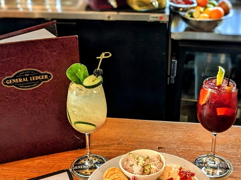 A cozy bar snack plate with crackers, pimento cheese, pickles, onions, and a creamy tuna salad, paired with colorful cocktails. A fun and flavorful spread perfect for happy hour.