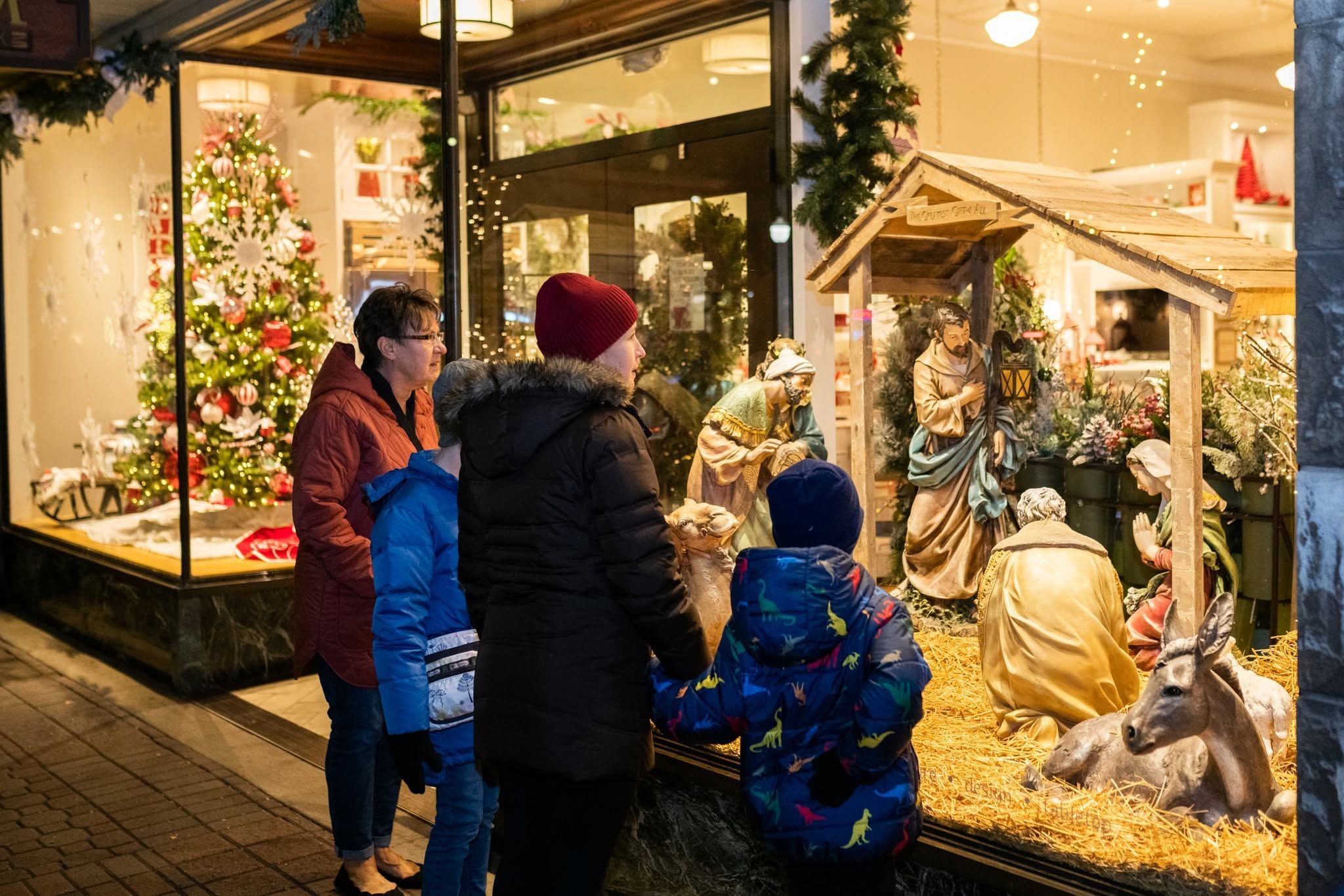 A family in winter coats pauses to admire a detailed outdoor nativity scene displayed beside a warmly lit holiday shop window during the Kerstmarkt Christmas market in Holland, Michigan.