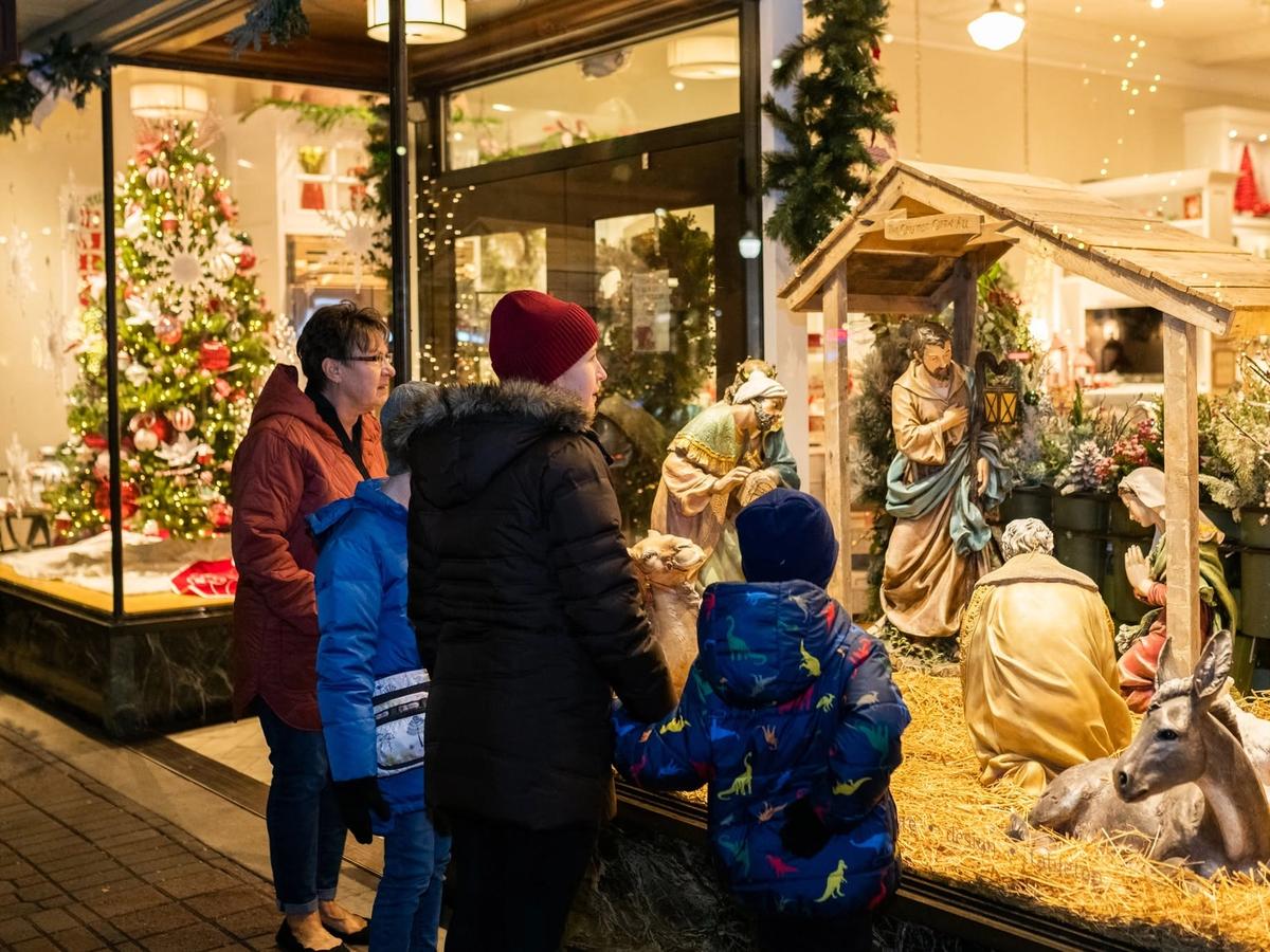 A family in winter coats pauses to admire a detailed outdoor nativity scene displayed beside a warmly lit holiday shop window during the Kerstmarkt Christmas market in Holland, Michigan.
