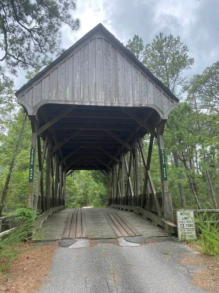A weathered gray wooden covered bridge with a peaked roof stretches across a wooded path, its interior trusses visible through the open entrance. "Private Road" signs flank both sides, and a posted weight limit sign stands to the right amid lush green trees under an overcast sky. 