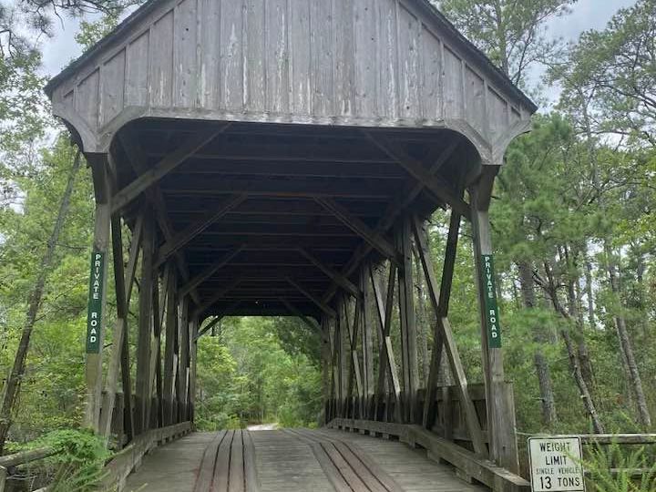 A weathered gray wooden covered bridge with a peaked roof stretches across a wooded path, its interior trusses visible through the open entrance. "Private Road" signs flank both sides, and a posted weight limit sign stands to the right amid lush green trees under an overcast sky.