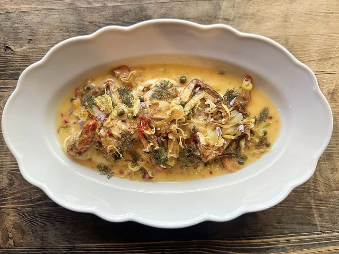 A scalloped white serving dish filled with a rich, golden seafood stew featuring soft-shell crab, capers, fennel, and herbs in a creamy, spiced broth, placed on a rustic wooden table.