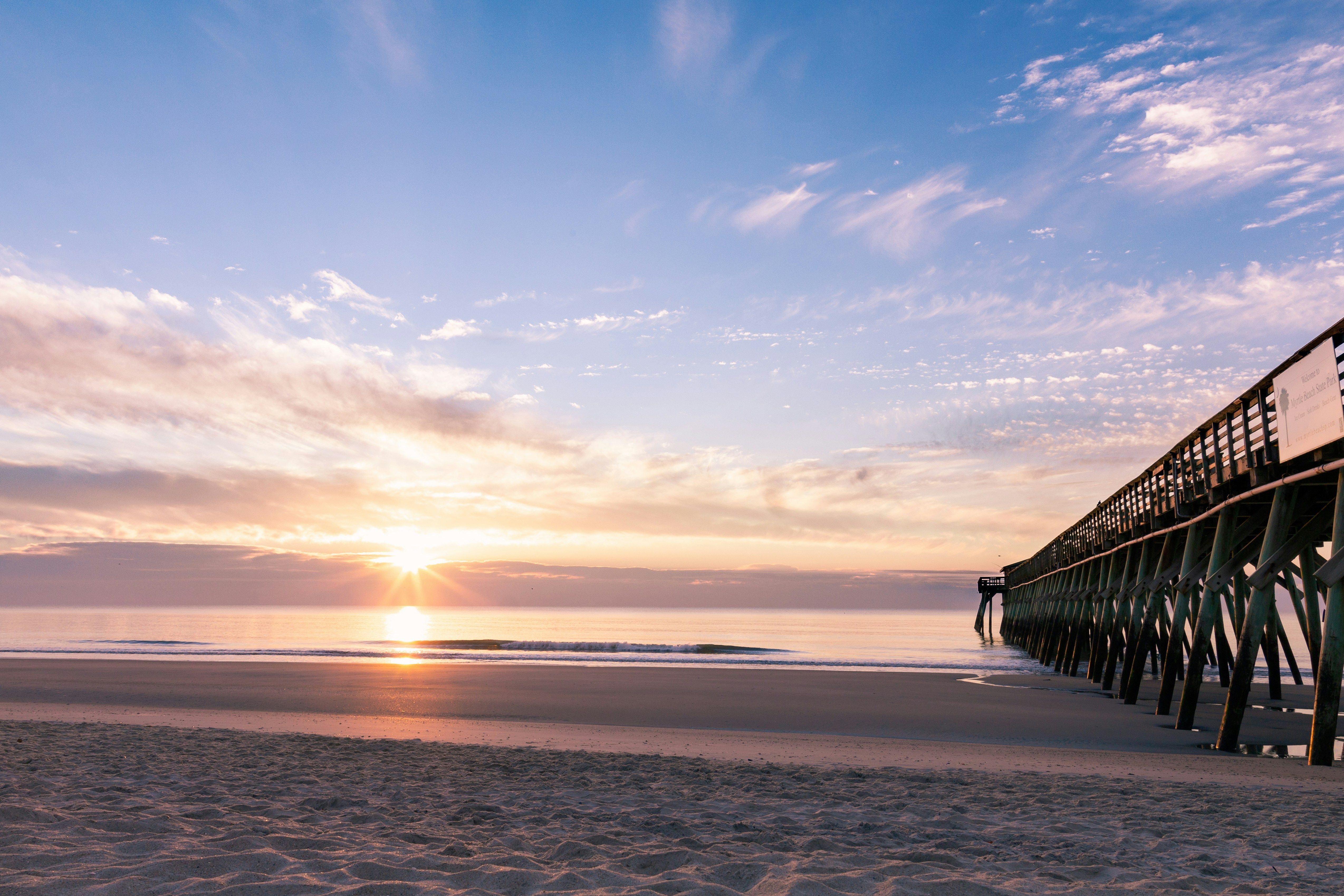 A calm beach sunrise with soft pastel clouds reflecting on the ocean, gentle waves washing the shore, and a long wooden pier extending from the right into the glowing horizon.