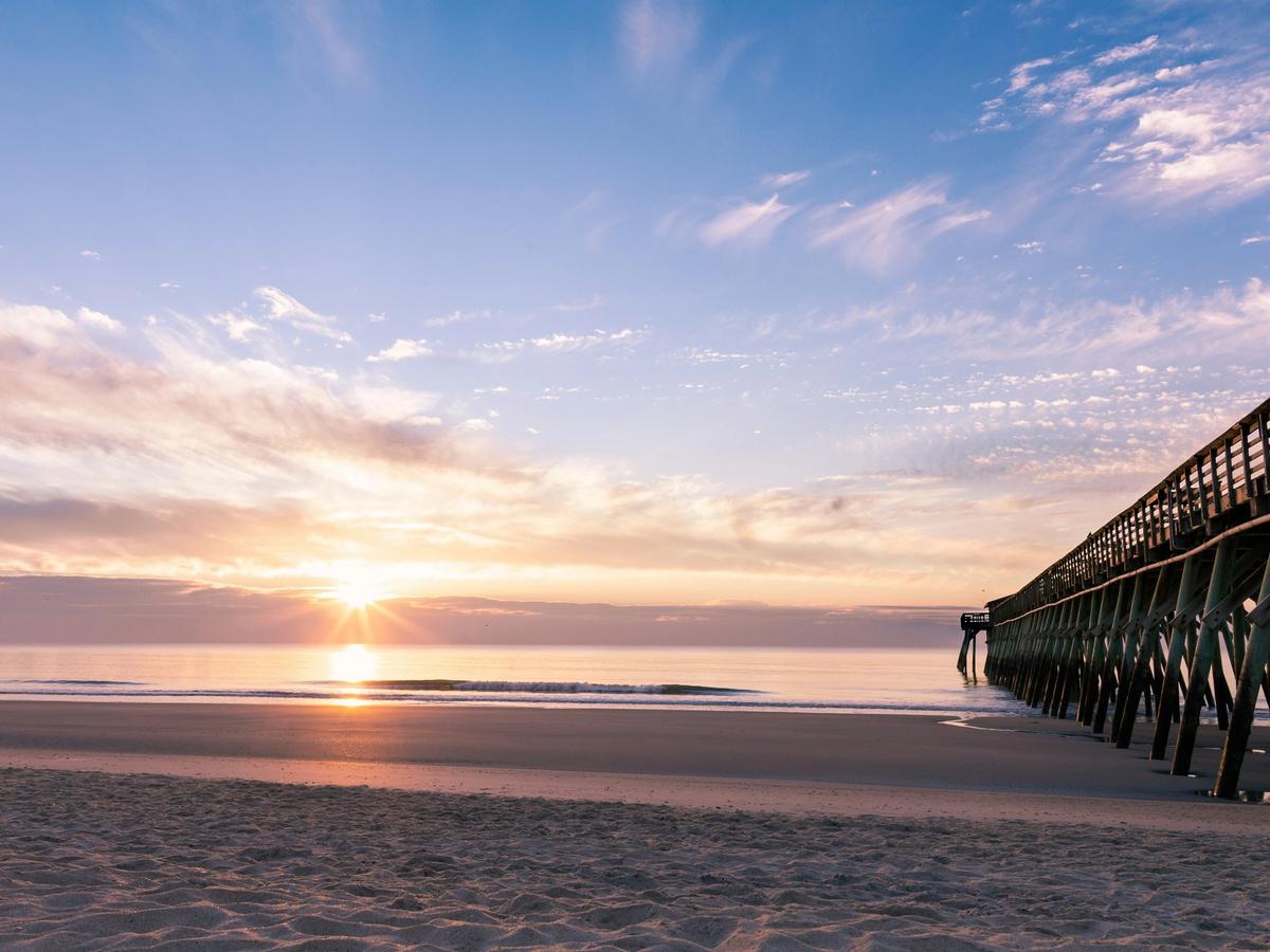 A calm beach sunrise with soft pastel clouds reflecting on the ocean, gentle waves washing the shore, and a long wooden pier extending from the right into the glowing horizon.