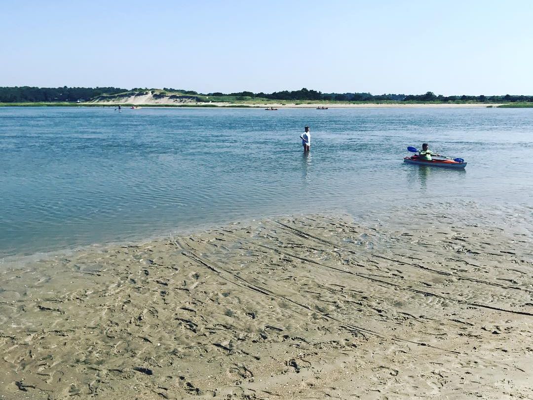 Alt Text: People enjoy kayaking and wading in shallow water near Mr. Beach Rentals along a quiet shoreline. The calm water makes it ideal for easy outdoor activities by the coast.