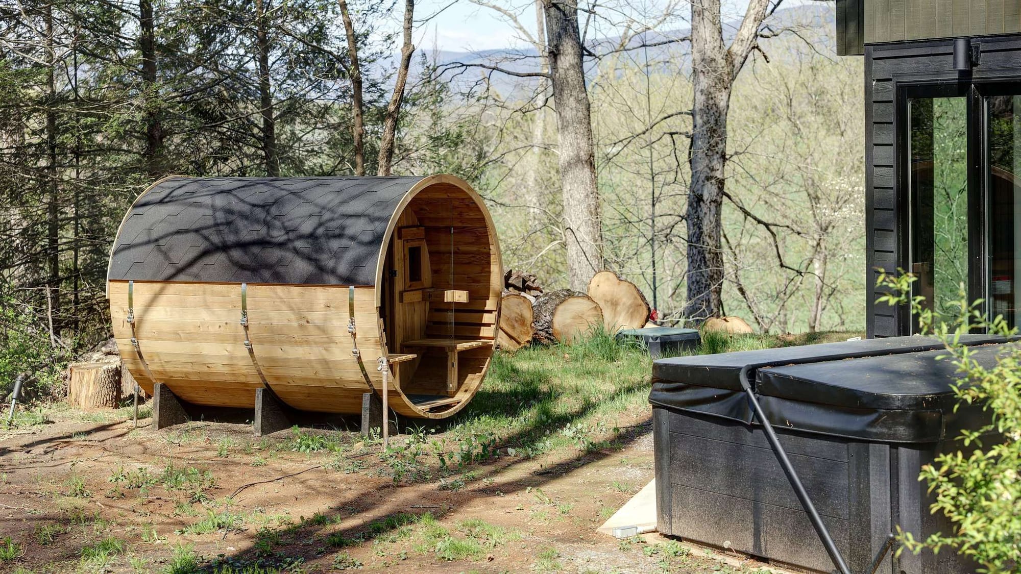 A cedar barrel sauna sits on a wooded hillside beside a covered hot tub, surrounded by bare spring trees with mountain ridges visible in the background. The rustic wellness setup is positioned just outside a modern black-framed building.