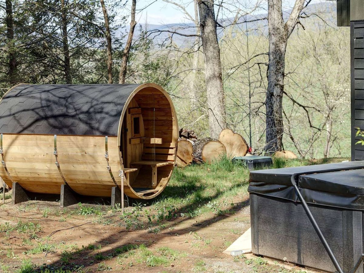 A cedar barrel sauna sits on a wooded hillside beside a covered hot tub, surrounded by bare spring trees with mountain ridges visible in the background. The rustic wellness setup is positioned just outside a modern black-framed building.