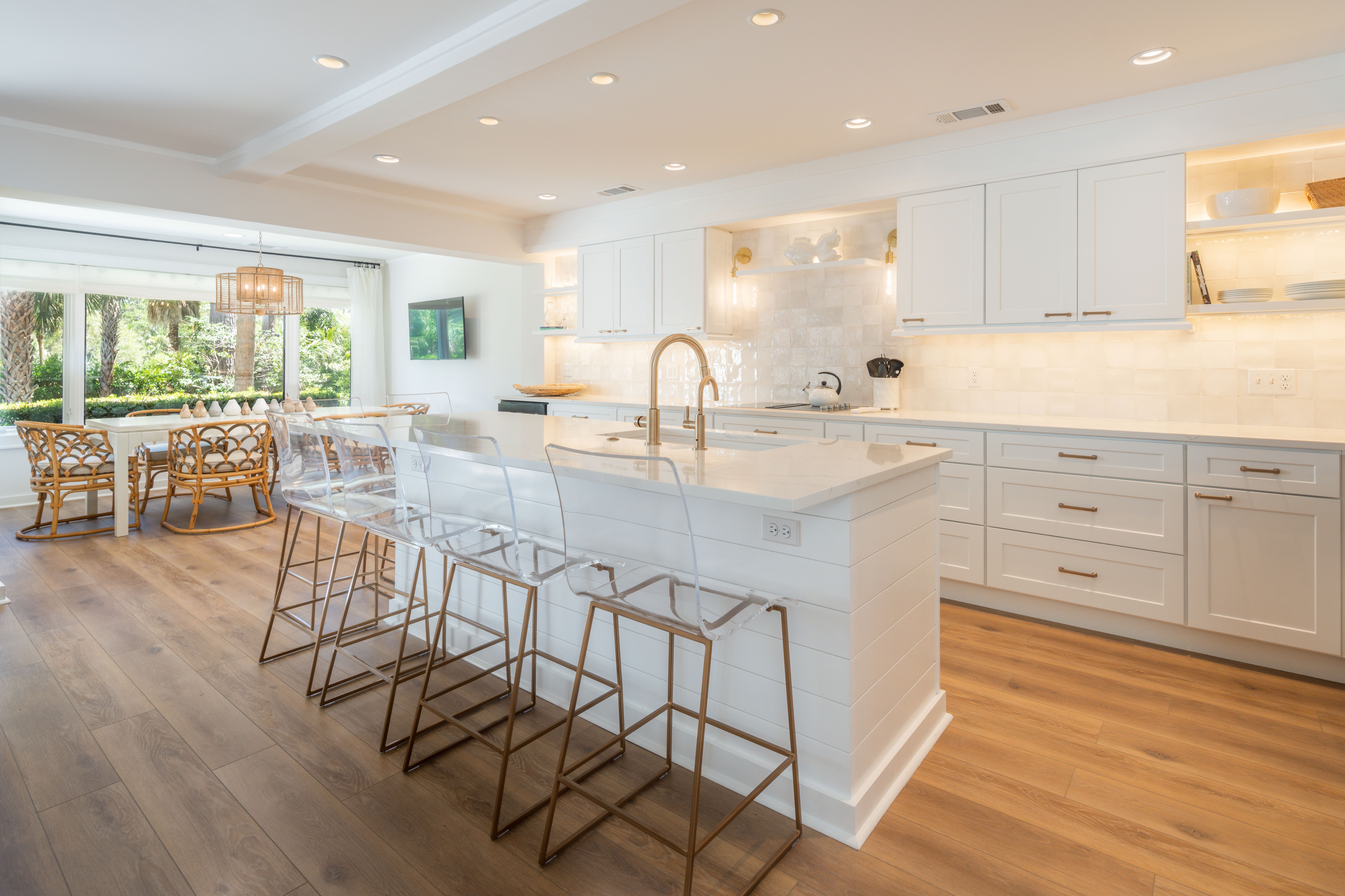 A bright, coastal-style kitchen features white cabinetry, a spacious island with a gold faucet, clear acrylic barstools with gold legs, and warm wood floors, leading into a sunlit dining area with rattan chairs and large windows overlooking palm trees.
