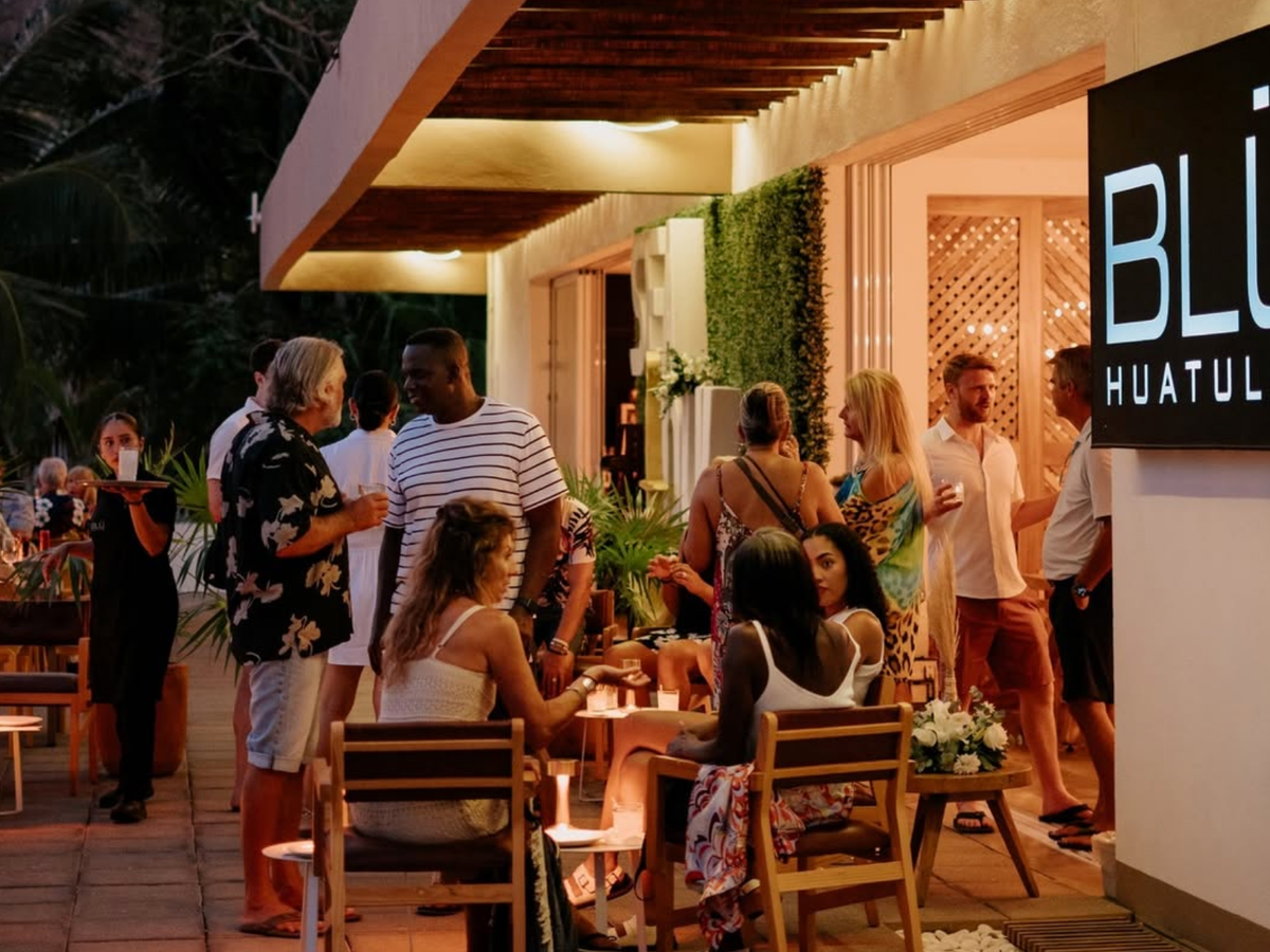 A lively evening scene outside BLÜ Huatulco, where people are mingling, chatting, and enjoying drinks in a stylish open-air setting with modern decor and warm lighting.