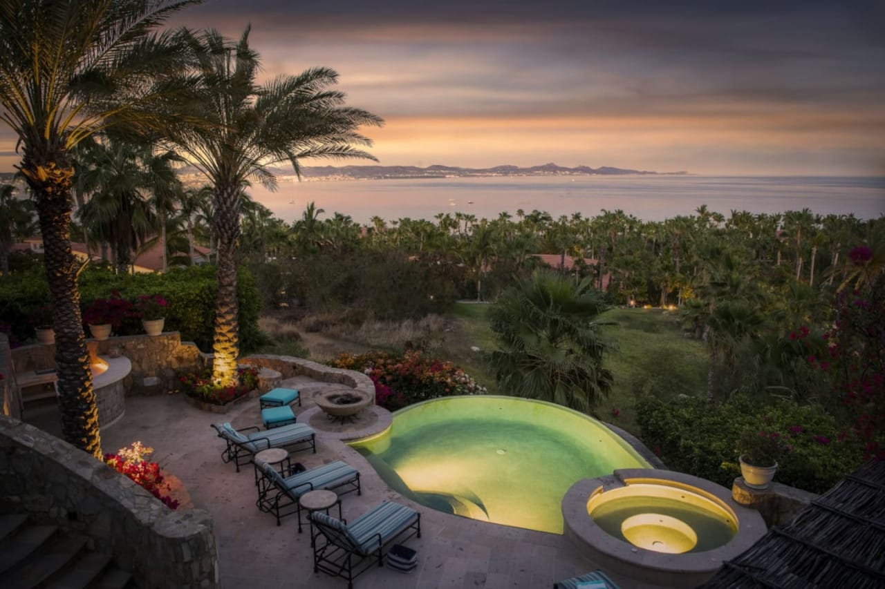 A glowing hillside pool sits under tall palm trees with a soft sunset over the ocean. Warm lights highlight the terraces and gardens, creating a dreamy, peaceful mood. The ocean views stretch all the way to the horizon, making the whole scene feel luxurious and calm.